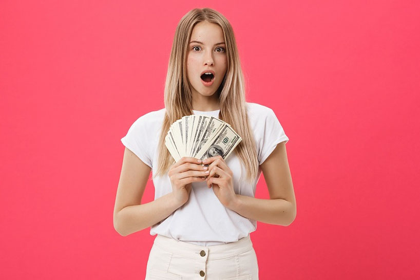 Young surprised shocked woman student with opened mouth holding bundle lots of dollars, cash money isolated on pink background. Education in high school university college