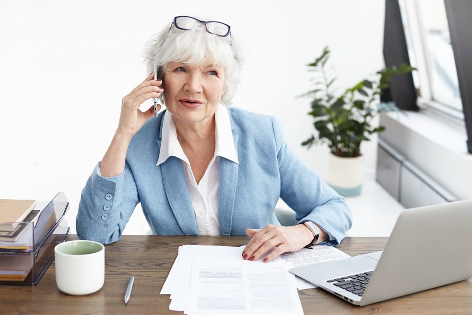 Beautiful mature woman with gray hair making phone calls in her office, Elegant senior female entrepreneur in stylish suit talking on mobile to potential partner, sitting at workplace with laptop